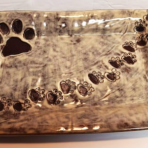 May include: A rectangular ceramic tray with a mottled cream and dark brown glaze. The surface features a large embossed paw print in the upper left corner and a trail of smaller paw prints extending diagonally across the center to the top right.