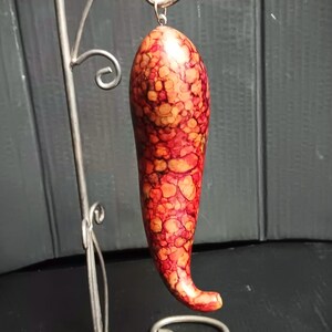 May include: A chili pepper-shaped ornament with a mottled red and orange pattern hanging from a silver-colored metal display stand. The stand features a curved hook at the top and a decorative base with multiple circular wire loops.