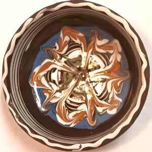 May include: A round ceramic bowl featuring a dark brown exterior with a white wavy rim pattern. The interior displays a marbled design with swirling patterns of white, orange, and light blue colors, creating a floral-like effect in the center.