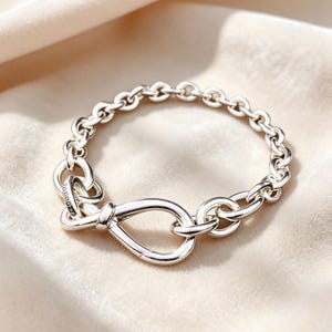 Può includere: Un bracciale a catena in argento lucido caratterizzato da una serie di maglie circolari interconnesse e da una maglia centrale a forma di infinito. La superficie metallica è liscia e riflettente, con una piccola targhetta incisa.
