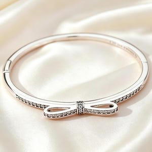 Könnte beinhalten: Ein Armreif in Roségold mit einem mittigen, schleifenförmigen Element. Die Oberfläche der Schleife und die Seiten des Armreifs sind mit kleinen, klaren, funkelnden Steinen besetzt. Das Metall hat eine polierte Oberfläche und einen Klappverschluss.