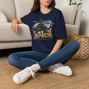 May include: A navy blue short-sleeve t-shirt featuring a graphic print of an AH-64 Apache attack helicopter in flight with a pilot visible in the cockpit. The word APACHE is printed in large, bold, textured tan letters below the helicopter.