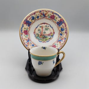 May include: A porcelain teacup and saucer set featuring a gold rim. The saucer displays a colorful landscape with a tower and floral motifs. The white teacup has a teal band, small blue floral accents, and a gold handle, resting on a black stand.