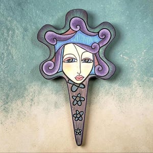 May include: A decorative wall plaque shaped like a stylized face with purple swirling hair, a blue crown-like top, and a pointed chin. The face features almond-shaped eyes, pink lips, and a lower section decorated with three small gray flower icons.