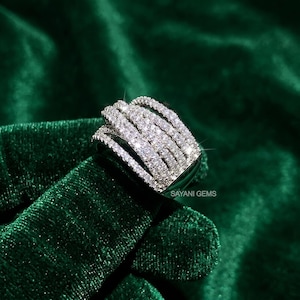 May include: A wide silver-toned ring featuring five parallel bands encrusted with numerous small, clear, sparkling stones. The ring displays the text SAYANI GEMS on its side, showcasing a multi-row design that creates a brilliant, reflective surface.