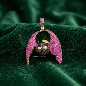 Könnte beinhalten: Ein fledermausförmiger Anhänger mit einem mit schwarzen Steinen besetzten Kopf, einem goldenen Heiligenschein und zwei weißen Steinaugen. Die Flügel sind mit rosa Steinen bedeckt, ebenso die Öse. Der Text SAYANI GEMS ist sichtbar.