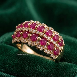 May include: A gold band ring featuring two horizontal rows of round, deep red rubies alternating with rows of small, clear white diamonds. The ring has a scalloped edge design and includes the text SAYANI GEMS printed on the side of the band.