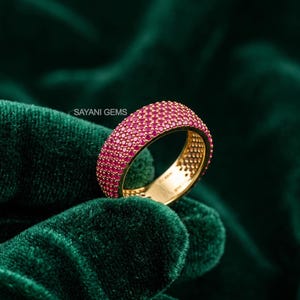 May include: A gold band ring featuring a pavé setting of numerous small, vibrant pink gemstones covering the entire outer surface. The interior of the band is smooth gold with a visible hallmark, and the text SAYANI GEMS is printed nearby.