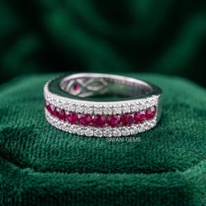 May include: A silver-toned band ring featuring a central row of round-cut red rubies flanked by two rows of small, clear, round-cut white stones. The metal band has a polished finish and the text SAYANI GEMS is visible on the inner surface.