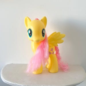 May include: A yellow plastic pony figurine with large blue eyes, a long pink mane, and a pink tail. The pony features molded wings on its back and a small butterfly symbol printed on its rear flank. The toy has a smooth, matte finish.