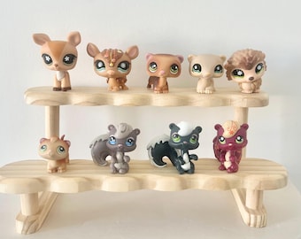 May include: A collection of nine small plastic animal figurines with large eyes, arranged on a two-tiered light wood display stand. The figures include a deer, a patterned creature, a ferret, a bear, a hedgehog, and several squirrels.