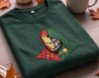 Può includere: Una felpa girocollo verde scuro con un ricamo dettagliato sul petto. Il disegno raffigura un personaggio dal volto diviso, con un lato che mostra una maschera rossa e dorata e l'altro una maschera verde metallizzata con cappuccio.
