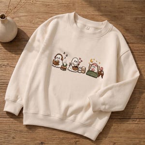 Puede incluir: Una sudadera de cuello redondo color crema con un estampado gráfico de tres fantasmas blancos. Un fantasma usa auriculares mientras lee, otro hornea con un cuenco y el tercero duerme en una cama con un antifaz rosa y una lámpara de noche.
