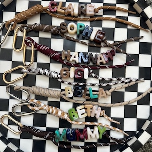 Può includere: Sette portachiavi realizzati con cordini in nylon intrecciato di vari colori, ciascuno dotato di un moschettone in metallo dorato o argentato e di perline con lettere in acrilico marmorizzato che compongono i nomi CLAIRE, SOPHIE, HANNAH, GIGI, BELLA, BETH e MUMMY.