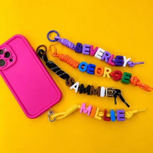Könnte beinhalten: Eine leuchtend pinke Smartphone-Hülle neben vier bunten Perlenbändern. Jedes Band verfügt über einen Metallring und Kunststoff-Buchstabenperlen, die die Namen BEVERLEY, GEORGE, SAMMIE und MILLIE bilden, befestigt an geflochtenen Kordeln in Lila, Orange, Schwarz und Gelb.