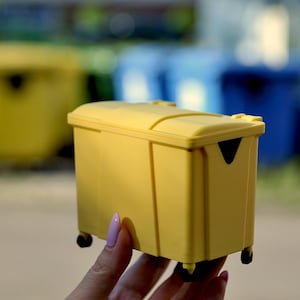Op de afbeelding: Een miniatuur gele plastic afvalbak met een scharnierend deksel en vier kleine zwarte wieltjes aan de onderkant. De kleine container heeft een rechthoekige vorm met verticale structurele ribbels en een zwarte driehoekige uitsparing.