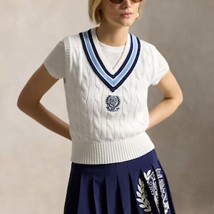 May include: A white cable-knit sweater vest with a V-neck featuring navy and light blue stripes, paired with a navy pleated skirt. The vest has a central embroidered crest, and the skirt displays a white graphic print with laurel leaf motifs.