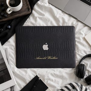 May include: A black laptop cover with a textured crocodile skin pattern, featuring a silver Apple logo in the center and the name Arnold Walker engraved in gold script lettering below the logo, designed for a portable computer device.
