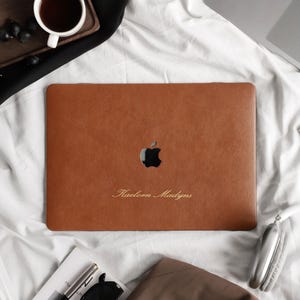 Peut inclure: Une protection d'ordinateur portable en cuir beige avec une découpe pour le logo Apple et le nom Kaelorn Madyns gravé en écriture cursive. La protection est appliquée sur la surface supérieure d'un ordinateur portable rectangulaire.
