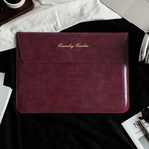 May include: A rectangular burgundy laptop sleeve with a textured crocodile skin pattern and a flap closure. The name Quinley Carlen is embossed in gold lettering on the front flap of the sleeve, which features a slim, flat, and structured design.