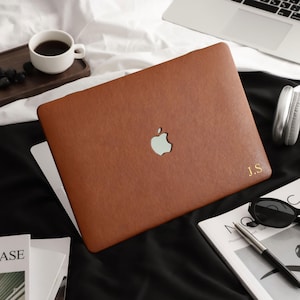 Puede incluir: Una funda para portátil de color marrón con un recorte central del logotipo de Apple en color plata y las iniciales J.S. grabadas en oro en la esquina inferior derecha. La funda tiene una textura lisa y mate para el exterior del equipo.