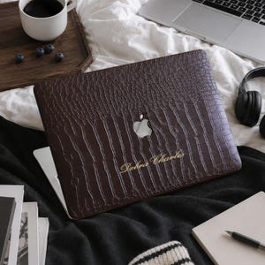 Könnte beinhalten: Eine dunkelbraune Laptop-Hülle mit einer strukturierten Krokodilleder-Optik und einem Ausschnitt für das Apple-Logo. Die Hülle ist im unteren mittleren Bereich mit dem goldfarbenen, kursiven Schriftzug Debra Charles versehen.