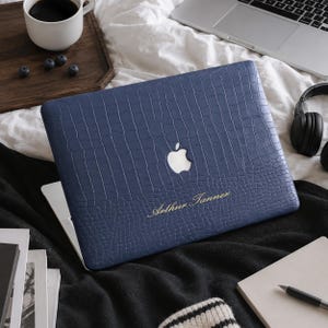 May include: A navy blue laptop case with a textured crocodile skin pattern. The case features a cutout for the Apple logo and the gold-colored text "Arthur Tanner" printed on the lower center of the cover, providing a protective skin.