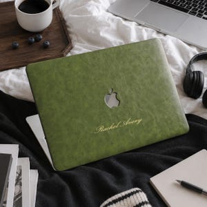 May include: A sage green faux leather laptop cover with a textured finish, featuring a cutout for the Apple logo and the name Rachel Avery engraved in a gold script font on the center of the cover, designed for a laptop computer.