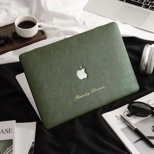 May include: A dark olive green laptop skin with a textured finish applied to a laptop. The skin features a cutout for the Apple logo and the gold-coloured engraved text "Brinley Dorian" positioned below the logo on the lower right side.