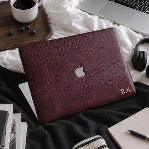 May include: A dark burgundy laptop skin with a textured crocodile pattern and a cutout for the Apple logo. The skin features the gold-colored initials R.K. printed in the bottom right corner, providing a custom look for the device.