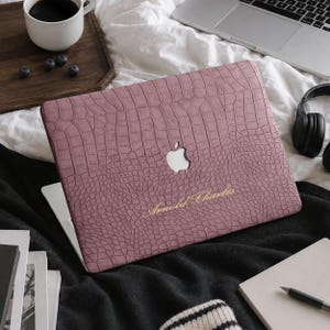 May include: A dusty rose laptop cover with a textured crocodile skin pattern, featuring a central cutout for the Apple logo and the gold-coloured cursive text "Arnold Charles" printed on the lower right side of the protective case.