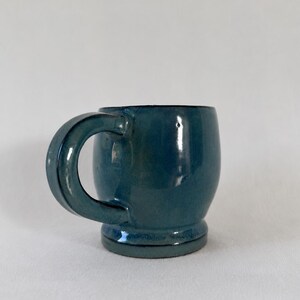 May include: A rounded ceramic mug with a glossy, deep teal blue glaze. The mug features a thick, curved handle on the side and a distinct, slightly flared circular base. The surface has a smooth, reflective finish with subtle variations in color.
