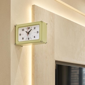 Op de afbeelding: Een rechthoekige wandklok met een lichtgroene omlijsting en een witte wijzerplaat. De klok is voorzien van zwarte Arabische cijfers voor 2, 3, 4, 6, 8, 9, 10 en 12, samen met zwarte uur- en minuutwijzers en een dunne rode secondewijzer.