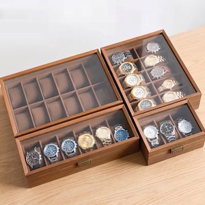 Puede incluir: Tres cajas de madera para relojes con tapas de cristal y compartimentos interiores marrones. Una caja grande vacía con doce ranuras, una mediana con seis relojes y una pequeña con tres relojes, todas con cierres metálicos.