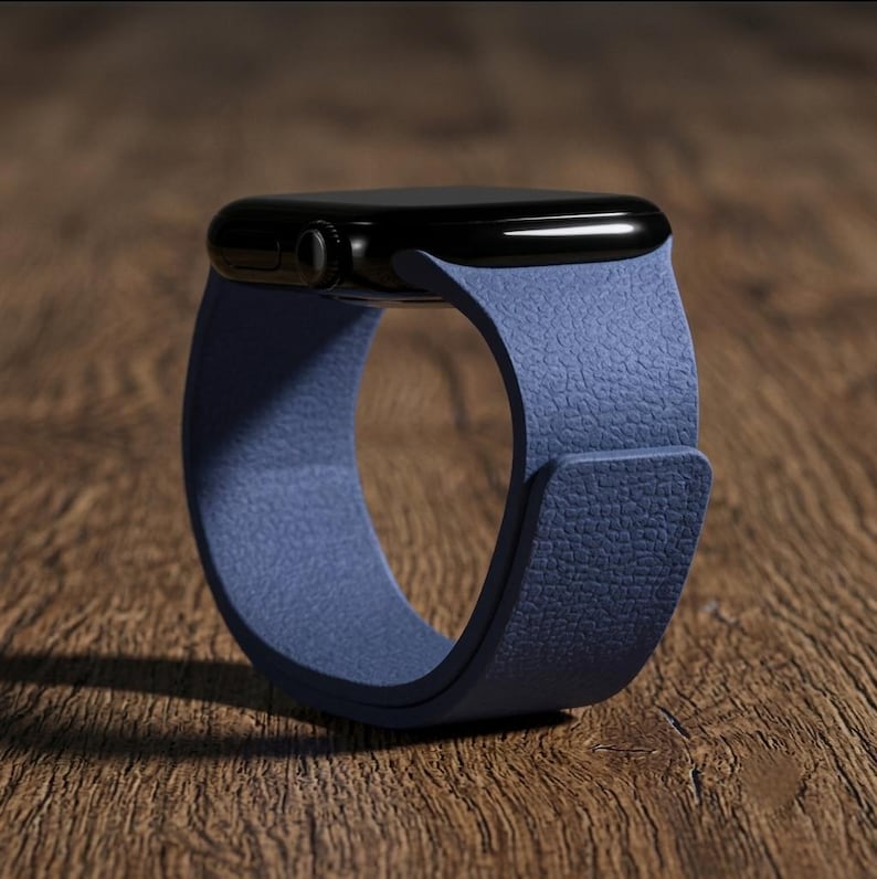 May include: A smartwatch with a black rectangular face and a textured blue leather band. The watch features a side crown and a smooth, rounded casing. The band has a visible grain pattern and a simple, clean design that wraps around the device.