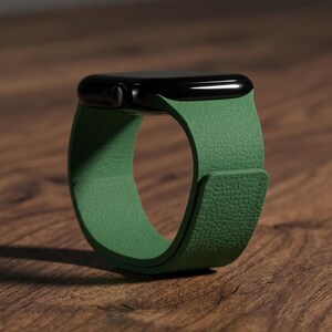 May include: A smartwatch with a black rectangular case and a textured green silicone band. The band features a smooth, matte finish with a distinct pebbled grain pattern, wrapping around to form a circular shape with a secure overlapping closure.