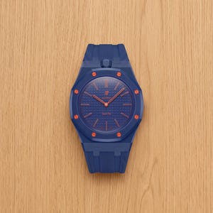 Könnte beinhalten: Eine blaue Armbanduhr mit strukturiertem Zifferblatt und orangefarbenen Akzenten. Die Uhr hat ein blaues Armband und ein gemustertes Zifferblatt mit orangefarbenen Zeigern und Markierungen. Auf dem Zifferblatt steht "AP" und "Royal Oak".