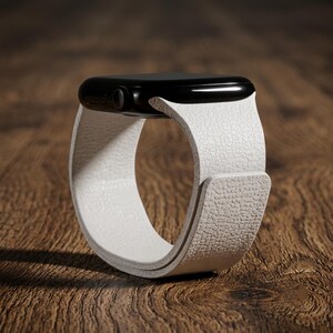 May include: A smartwatch featuring a glossy black rectangular case and a textured white strap. The strap has a pebbled surface finish and wraps around to form a circular loop. The side of the watch case displays a small circular digital crown button.