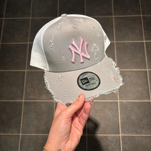 Pode incluir: Um boné trucker cinzento e branco com aba e painel frontal desgastados e desfiados. O boné apresenta um logótipo dos New York Yankees bordado em rosa na frente e um autocolante circular preto da New Era na aba com a palavra Adjustable.