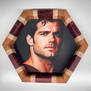 May include: A hexagonal wooden frame featuring alternating segments of dark purple and light tan wood. The centre contains a portrait of a man with dark hair, blue eyes, and light stubble, wearing a dark red textured garment against a dark background.