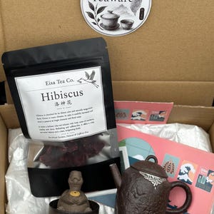 Peut inclure: Une boîte cadeau en carton contenant un sachet noir de thé Hibiscus Eisa Tea Co., une théière texturée marron foncé, une petite tasse assortie avec une figurine de tortue, un autocollant Taiwan Teaware et une carte rose illustrée.