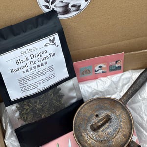 May include: A cardboard box containing a black pouch of Black Dragon Roasted Tie Guan Yin tea with a white label, a round metallic brown teapot with a long handle, a circular Taiwan Teaware sticker, and small decorative cards with illustrations.