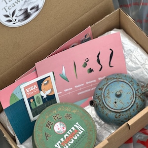 May include: A cardboard box containing a blue speckled ceramic teapot, a round green tin labelled Tuocha with a crane illustration, a pink informational card with tea types, and a small square Eisa Tea Co card with a crane and leaf graphic.