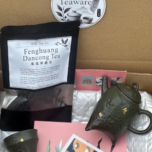 May include: A cardboard box containing a black pouch of Fenghuang Dancong Tea, a dark green textured teapot with gold accents, a matching tea cup, a circular Taiwan Teaware sticker, and a pink Eisa Tea Co. card with a graphic illustration.