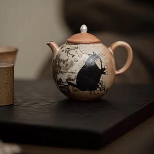 Pode incluir: Um bule de cerâmica redondo com acabamento em tom bronzeado e castanho manchado, apresentando uma ilustração de um gato preto na lateral. O bule tem uma tampa a condizer com um pequeno botão branco, uma pega curva e um bico curto e texturado.