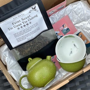 May include: A cardboard box containing a black pouch of Eisa Tea Co. Faint Scent Tie Guan Yin Oolong tea, a textured olive green teapot with a lid, and two matching olive green tea bowls featuring small black fish illustrations inside.