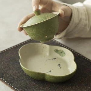 May include: A green ceramic gaiwan with a textured finish and a matching lid, paired with an oval-shaped, off-white ceramic saucer featuring a green lotus leaf illustration and small black fish motifs painted on the interior surface.