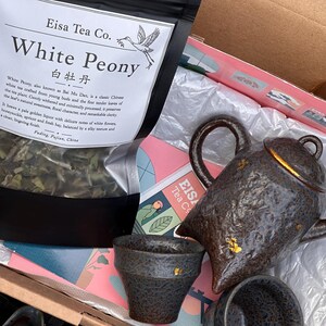 May include: A gift box containing a bag of Eisa Tea Co. White Peony tea with a label featuring a crane illustration, a dark brown textured ceramic teapot with gold accents, and matching ceramic tea cups nestled in white tissue paper.