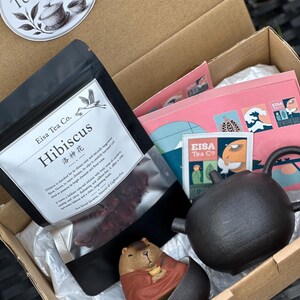 May include: A cardboard gift box containing a black pouch of Eisa Tea Co. Hibiscus tea, a black textured teapot, and a small ceramic figurine of a capybara wearing a red robe inside a dark bowl. A Taiwan Teaware sticker is on the box lid.
