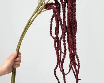 May include: A bundle of artificial amaranth stems featuring long, cascading, deep burgundy flower plumes with a textured, granular appearance. The stems are a muted olive green color, branching out to support the drooping, rope-like floral strands.